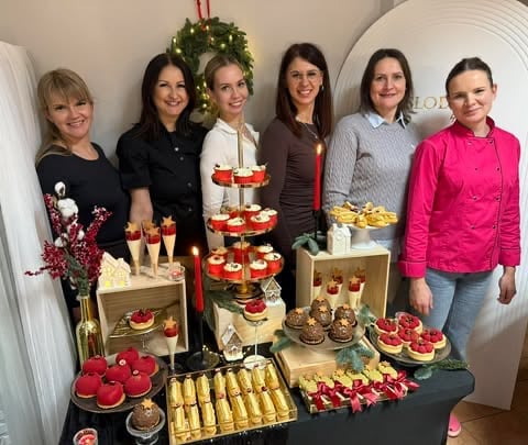 Six women standing behind a holiday dessert display with tiered cake, pastries, chocolates, and festive decorations