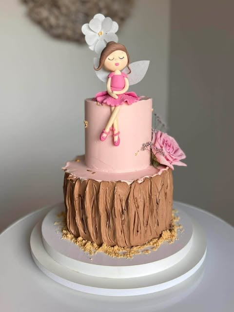 Tiered cake with brown wood texture bottom layer and pink top layer, topped with fondant fairy figurine with white flower hat