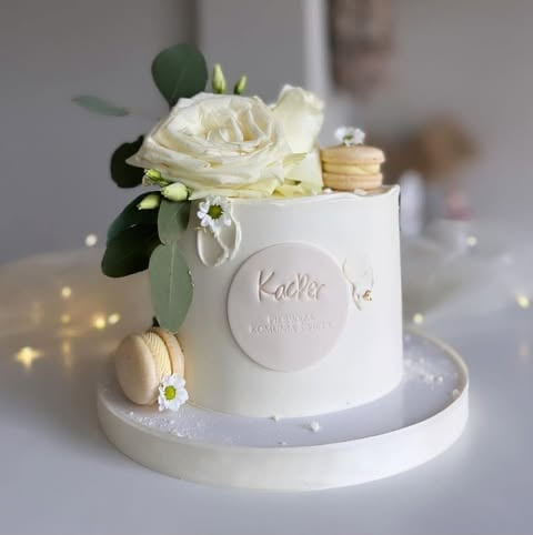 White tiered cake decorated with cream roses, eucalyptus leaves, macarons, and a circular Kacher logo plaque on front
