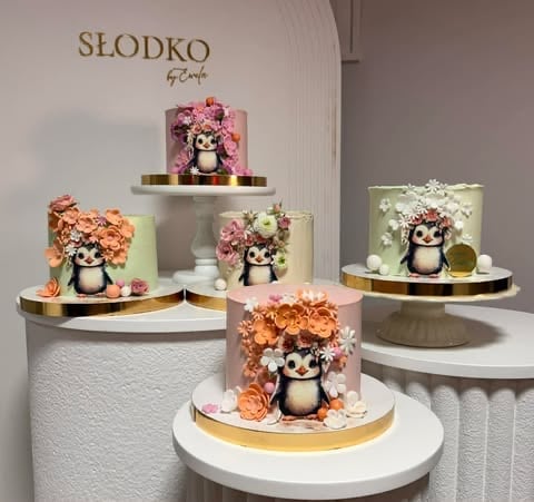 Collection of decorative penguin cakes with colorful flower arrangements on white and gold pedestals, branded SLODKO