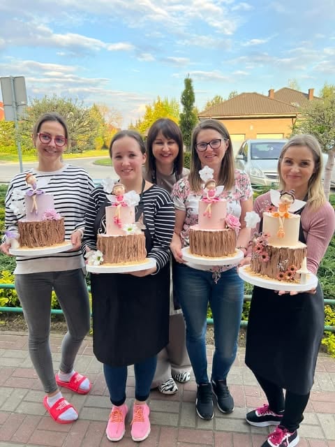 Five women holding decorated chocolate cakes with pink floral accents, standing outdoors in front of a house on a sunny day