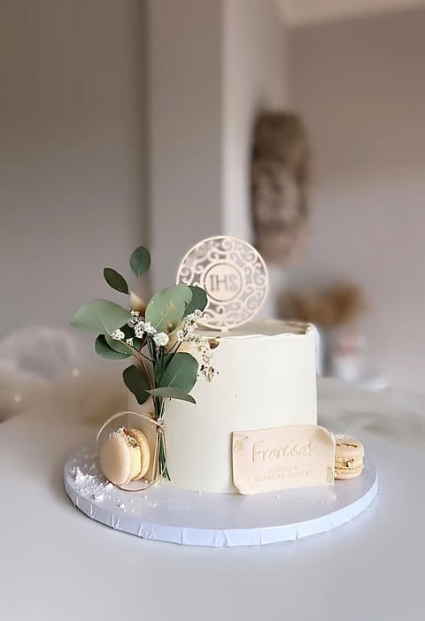 White wedding cake on pedestal stand with eucalyptus, gold accents, and decorative topper on neutral background