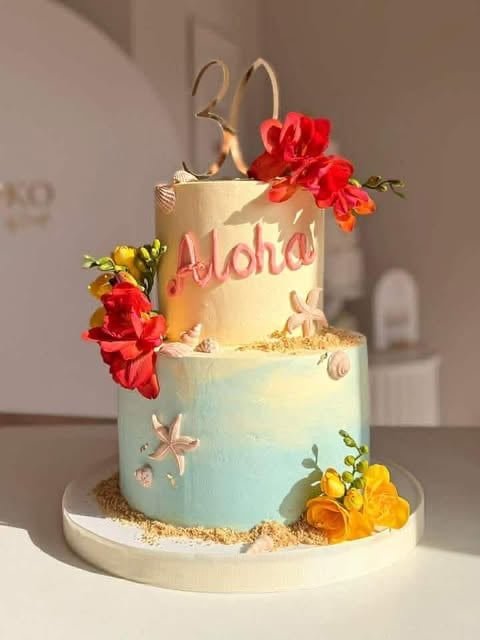 Two-tiered aloha-themed cake with cream and mint frosting, decorated with red and yellow flowers, starfish, and beach elements