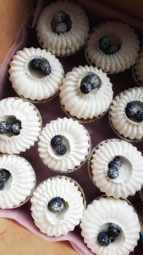 Multiple cream-colored tartlets topped with fresh blueberries arranged in a pink box