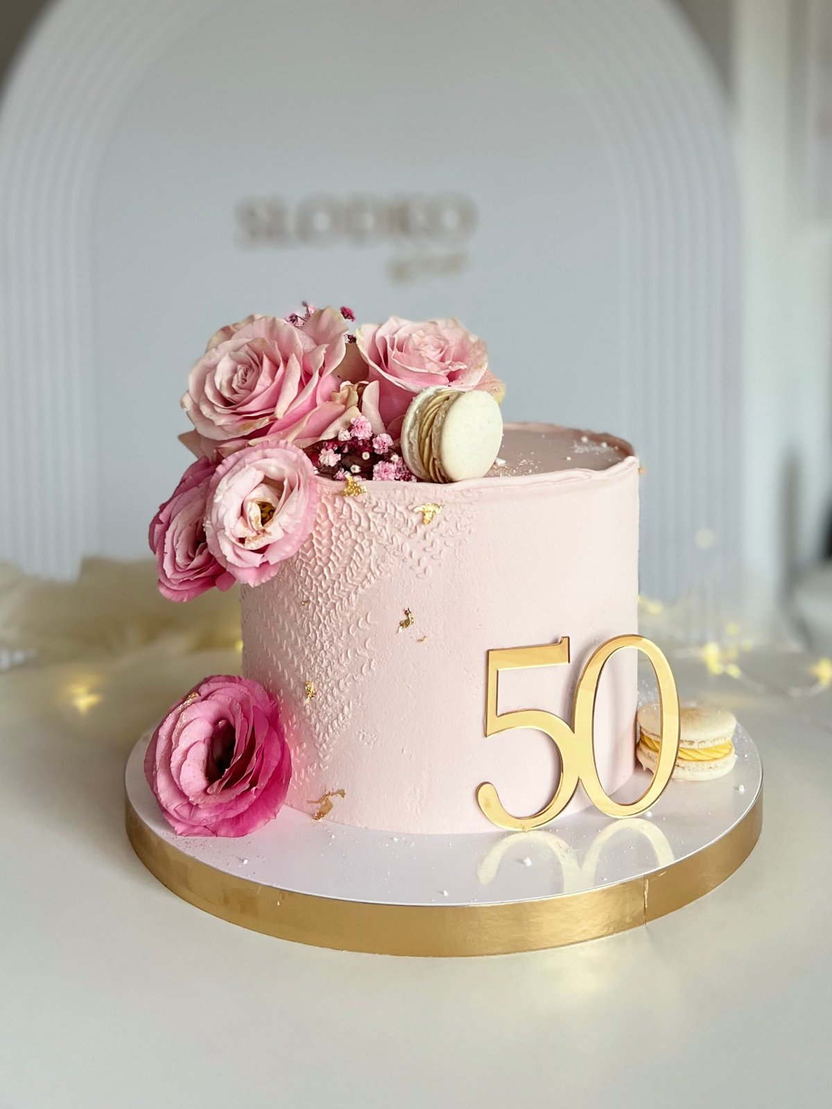 Elegant 50th birthday cake with pink textured frosting, fresh pink roses, gold number 50, macarons, and gold base