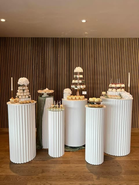 Elegant dessert display with white tiered cakes and macarons on fluted white pedestals against a dark striped wall