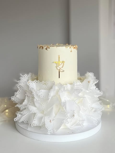 Two-tiered white wedding cake with gold cross decoration and white lace ruffles on white plate