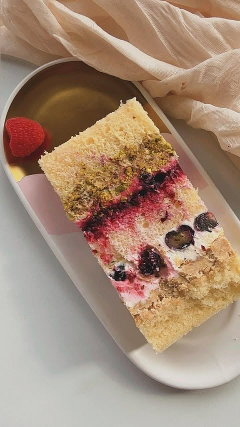Layered vanilla cake slice with pink frosting, berries, and pistachio crumble on a white plate with a fresh raspberry and drizzle