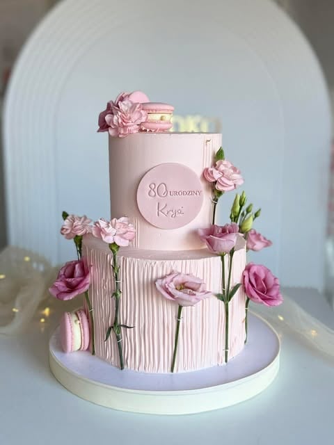 Two-tier pink birthday cake decorated with fresh pink carnations and the text 80 yaşındaysın Kgc, displayed on a white cake stand