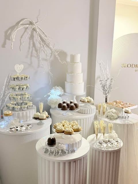 Elegant dessert display with tiered white cake, cupcakes, chocolates, and pastries arranged on white pedestals with gold accents and frosted branches