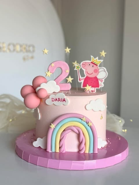 Pink birthday cake decorated with Peppa Pig character, number 2 topper, rainbow design, clouds, and gold stars on purple base