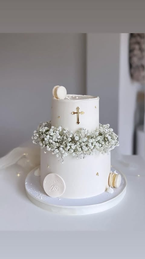 Two-tiered white cake decorated with a gold cross, babys breath flowers, macarons, and religious medallion on white cake stand