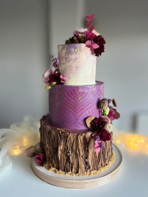 Three-tiered decorative cake with brown bark texture base, purple middle tier, and white top tier adorned with pink and burgundy flowers