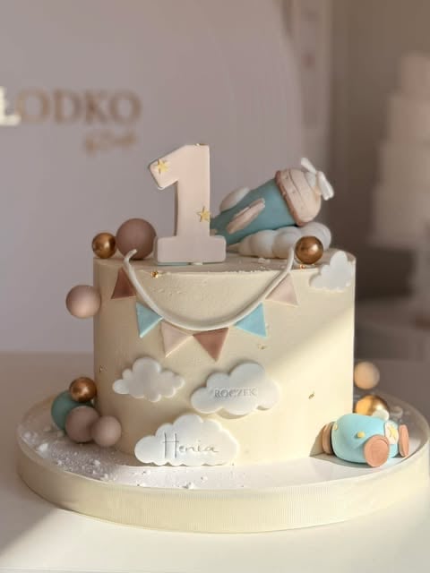 First birthday cake with cream frosting, number 1 topper, decorative balloons, clouds, bunting, and toy train figurine