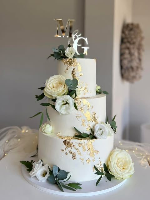 Three-tiered white wedding cake with gold leaf accents, fresh white roses, eucalyptus greenery, and Mr and Mrs gold topper