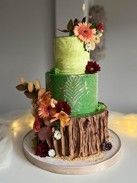 Three-tiered decorative cake with textured layers in green and brown, adorned with fresh coral and burgundy flowers and foliage