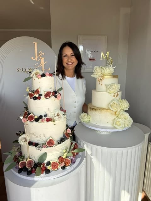 Woman standing between two ornate wedding cakes decorated with fresh figs, berries, and flowers, with custom cake toppers