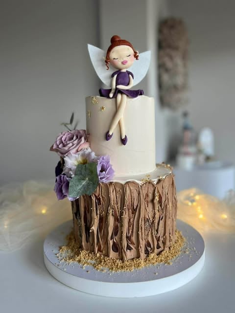 Decorative cake featuring a fairy figure with red hair and white wings on top, purple flowers, and textured chocolate bottom layer