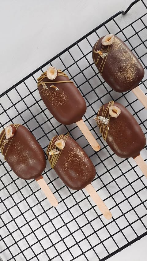 Five chocolate-covered ice cream bars with gold glitter and almond slices on a black grid napkin