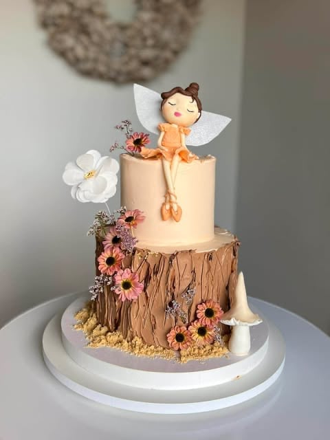 Whimsical fairy-themed tiered cake with winged character topper, floral decorations, wood-textured bottom tier, and sitting on a white plate