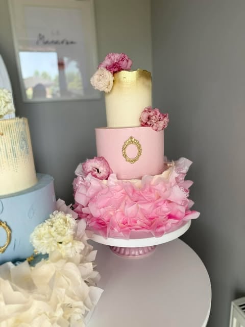 Three-tiered elegant wedding cake with pale yellow top tier, pink middle tier with gold oval detail, and ruffled pink bottom tier, decorated with pink and white carnations
