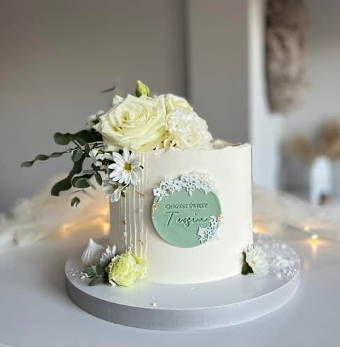 Elegant white celebration cake topped with cream roses, white flowers, and greenery on a white cake board with string lights