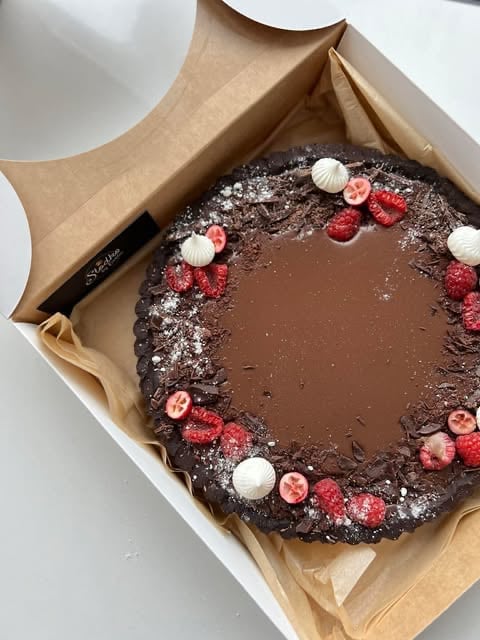 Round chocolate cake decorated with fresh raspberries, white pearls, and powdered sugar, placed on brown parchment paper in a white box