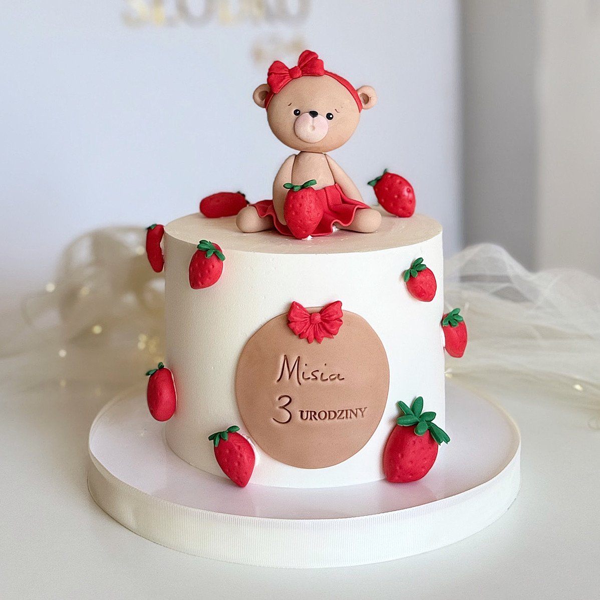 Strawberry-themed birthday cake with a cute bear topper wearing pink, decorated with red strawberries and a third birthday message