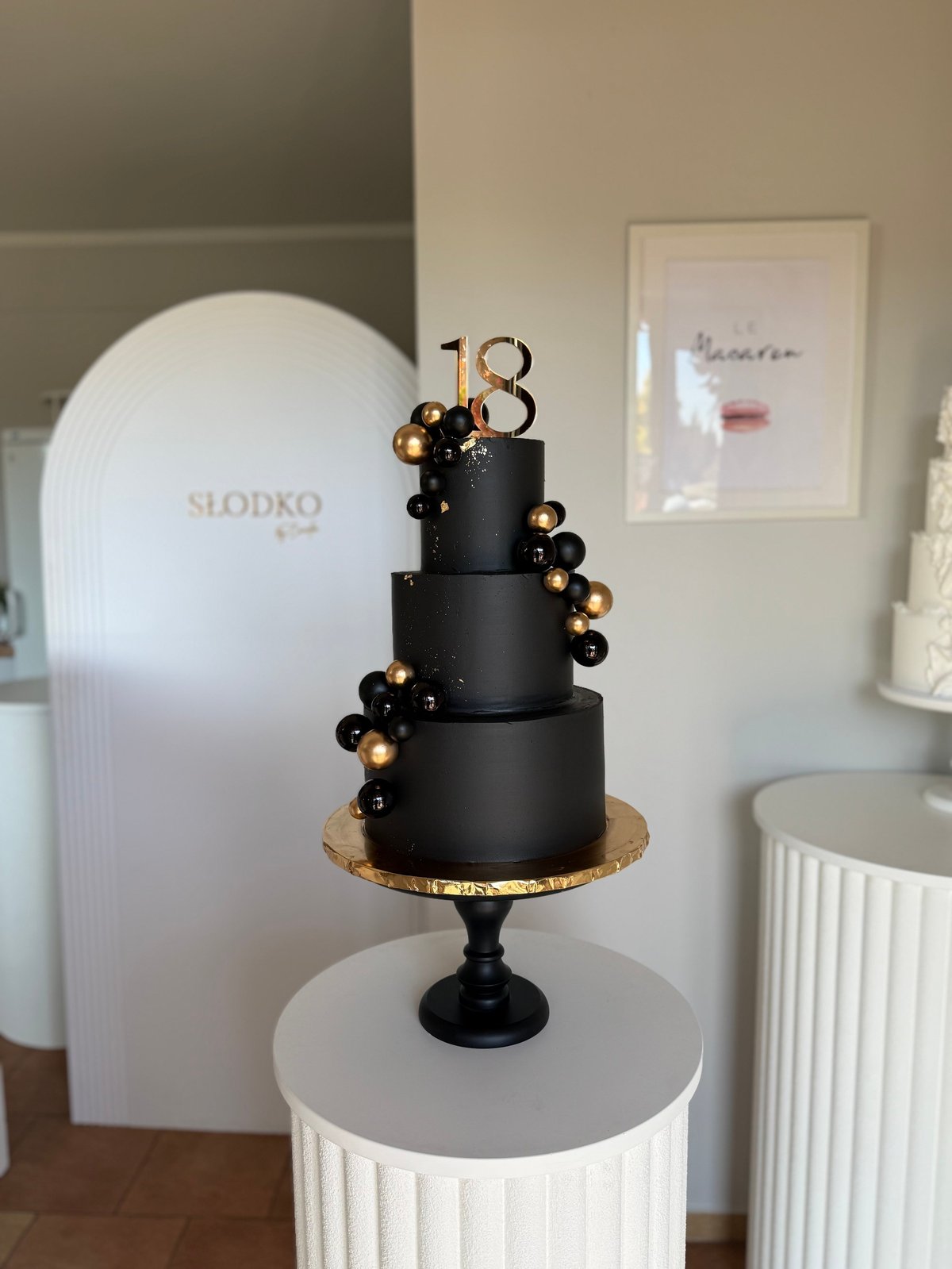 Elegant three-tiered black cake decorated with gold and black spheres and a gold number 18 topper on white pedestal display