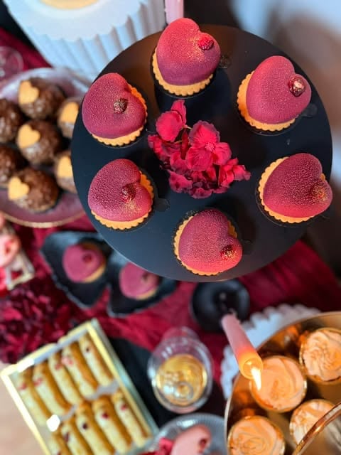 Six pink raspberry mousse domes with gold bases arranged on a black plate with fresh flowers