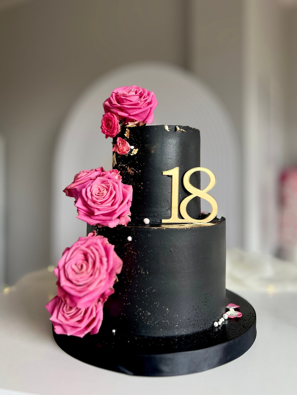 Two-tier black birthday cake decorated with pink roses and gold number 18, displayed on white surface