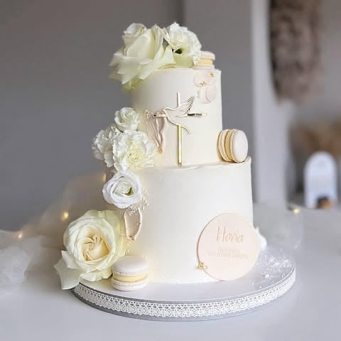 Elegant two-tier white wedding cake decorated with cream roses, gold accents, macarons, and a cross topper on a silver base