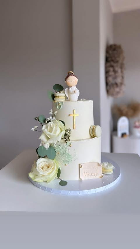 Two-tier white religious celebration cake with gold cross, white roses, greenery, and a figurine topper on a white stand