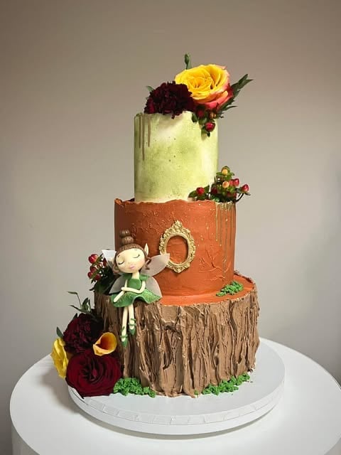 Three-tiered decorative cake with textured wood-grain base tier, terracotta middle tier, pale green top tier, adorned with colorful flowers and a small fairy figurine