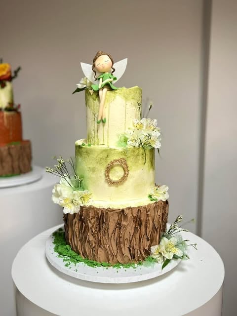 Three-tiered woodland-themed cake with green and yellow frosting, wood texture base, fairy figurine on top, and white flowers as decoration