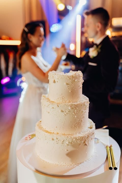 Three-tiered white wedding cake with gold sparkle decoration on white stand, couple dancing in blurred background at reception