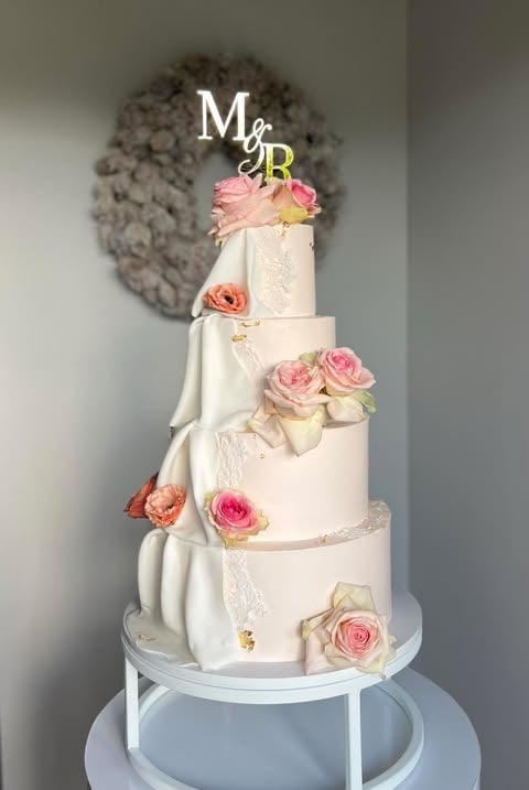 Elegant multi-tiered wedding cake with white frosting, fresh pink roses, and decorative draping, topped with Mr and Mrs initials