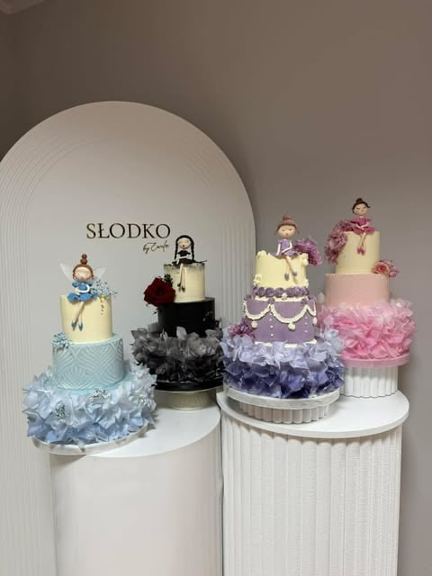 Display of four tiered wedding cakes with toppers, decorated ruffles, and displayed on white cylindrical pedestals against a textured wall