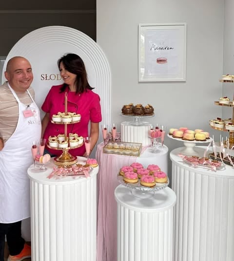 Man and woman standing in front of an elegant dessert display with pink and white decorated macarons, pastries, and tiered stands at a bakery or confectionery shop