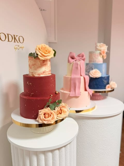 Display of three multi-tiered wedding cakes on white pedestals, featuring red, pink, and blue tiers decorated with roses and bows