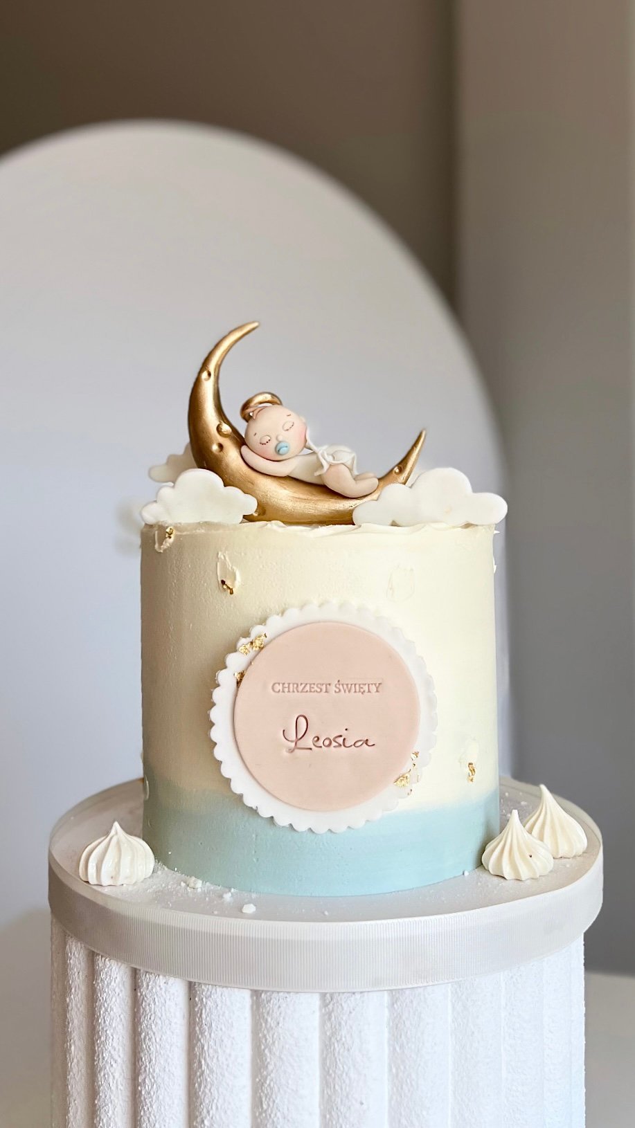 Elegant two-tiered cake with baby on golden crescent moon topper, personalized with name Lesia, clouds and meringue details