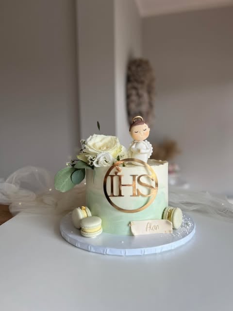A religious celebration cake with pale green frosting, IHS symbol, small figurine of a child, white flowers, macarons, and decorative elements on a white plate