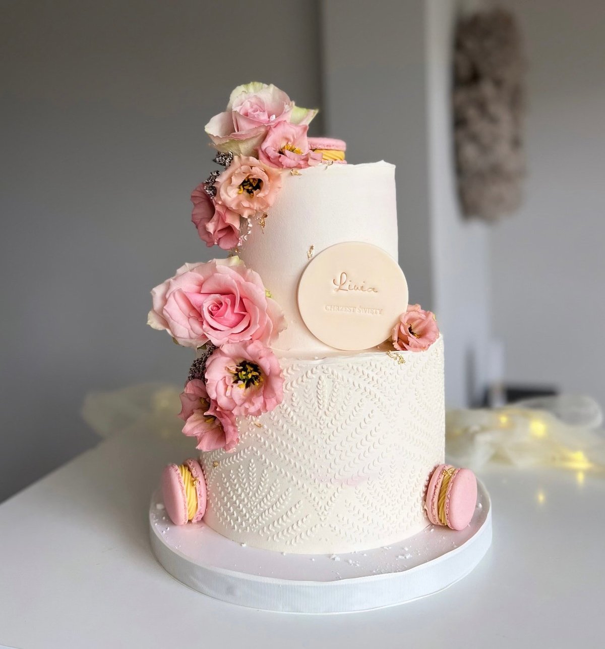 Three-tiered white wedding cake decorated with pink flowers, macarons, and gold accents on a white stand