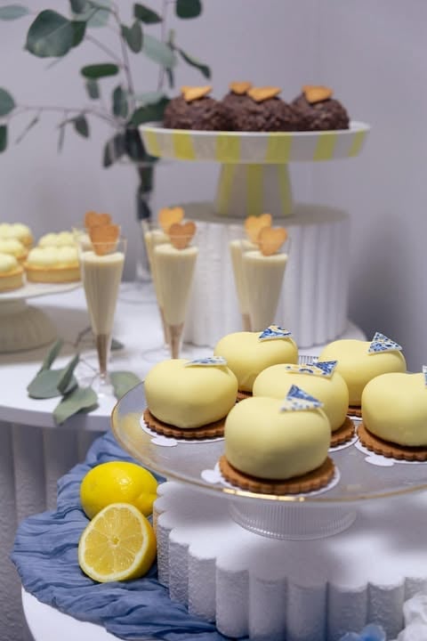 Elegant dessert display with yellow dome cakes, lemon slices, chocolate brownies, and cream desserts on white cake stands with eucalyptus decoration