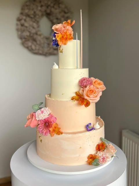 Four-tiered wedding cake with cream frosting, decorated with fresh pink, orange and purple flowers, metallic accents, and two tall white candles on top