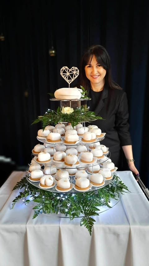Woman in black blazer smiling beside a tiered wedding dessert display with geometric heart topper and green foliage