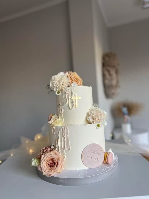 Two-tiered white wedding cake decorated with flowers, gold cross, and beaded details on white stand