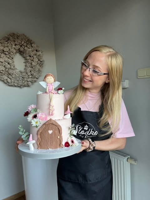 Young woman wearing glasses and black apron holding a decorative two-tier cake with flowers and a fairy figurine