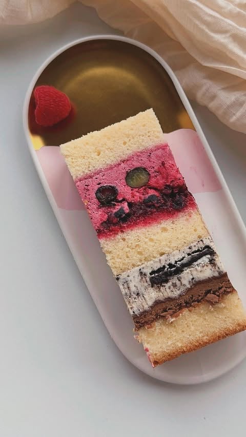 Multi-layered cake slice with vanilla, pink berry, and chocolate layers topped with a fresh raspberry on a pink plate