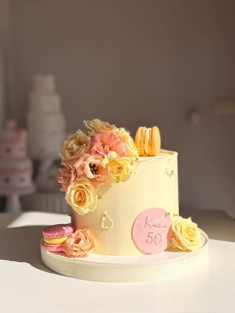 Cream-colored fondant cake decorated with fresh coral and yellow roses, macarons, and a pink circular disc reading 50 on a white cake board
